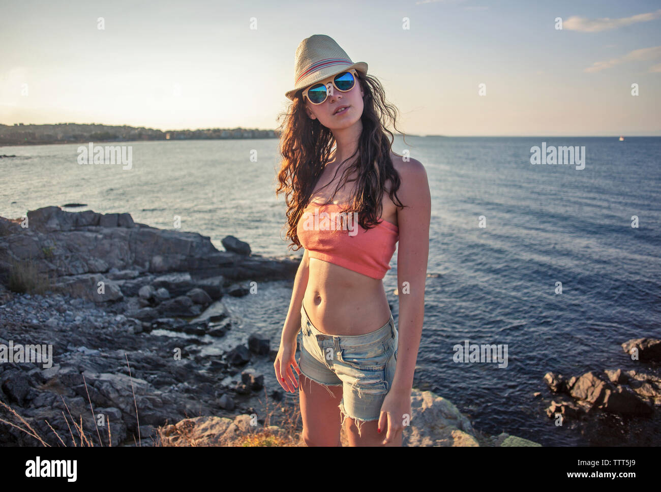 Ritratto di fiducioso giovane donna che indossa gli occhiali da sole e cappello mentre permanente sulla spiaggia rocciosa durante il tramonto Foto Stock