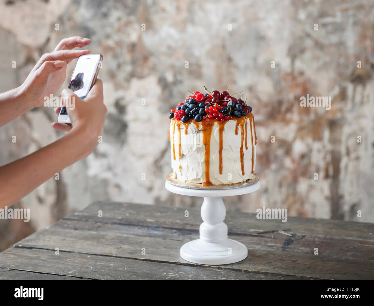 Tagliate le mani della donna fotografare la torta con un telefono cellulare sul tavolo di legno contro la parete arrugginito a casa Foto Stock