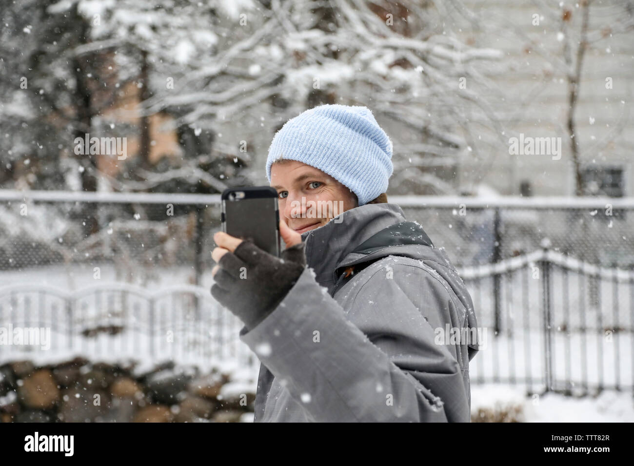 Uomo sorridente indossando un abbigliamento caldo durante l'assunzione di selfie con smart phone durante la nevicata Foto Stock