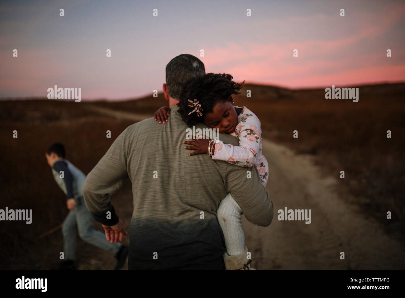 Vista posteriore del padre che porta la figlia mentre si cammina sulla strada sterrata durante il tramonto Foto Stock