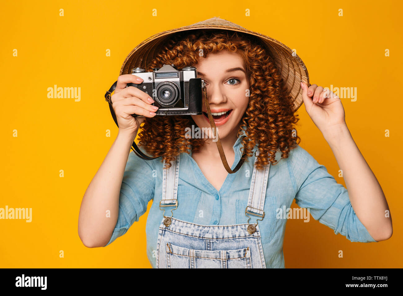 Ricci sorridente i capelli rossi donna che indossa cappello cinese azienda foto fotocamera e fare foto su sfondo giallo. Turismo e Viaggi Foto Stock