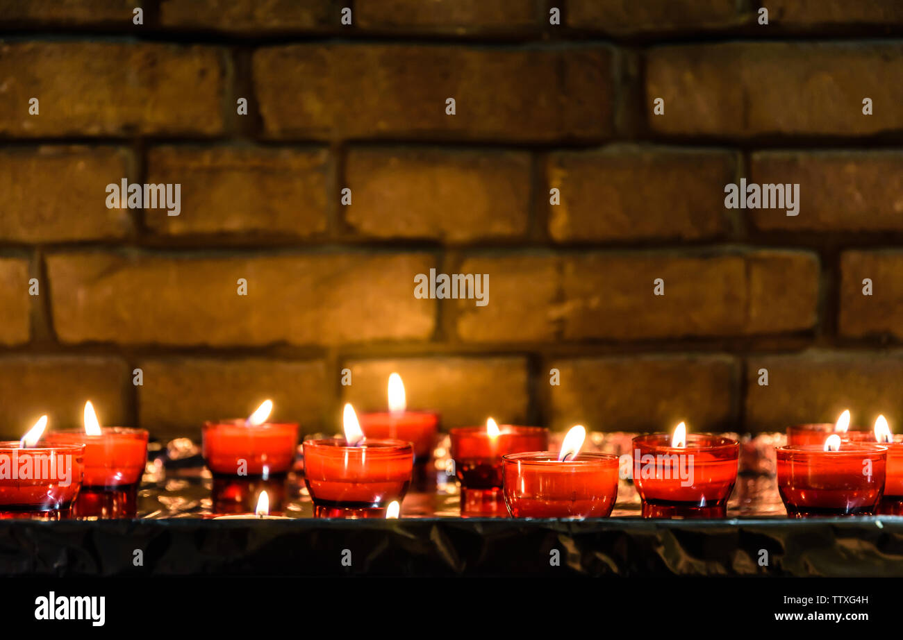 Bianco di piccole candele votive illuminato e posto in rosso i titolari di vetro su un tavolo ricoperto con un foglio di alluminio di fronte a un muro di mattoni in una chiesa. Foto Stock