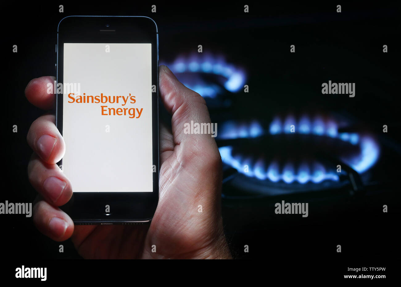 Un uomo che guarda il logo del sito web per l'energia azienda Sainsbury's energia sul suo telefono nella parte anteriore della sua cucina a gas (solo uso editoriale) Foto Stock