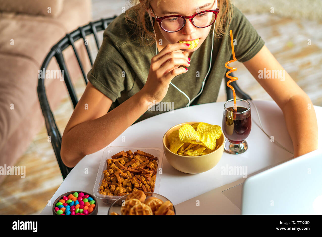 Ragazza lavora a un computer e si mangia il fast food. Cibo malsano: chips, cracker, candy, waffle, cola. Il cibo spazzatura, concetto. Foto Stock