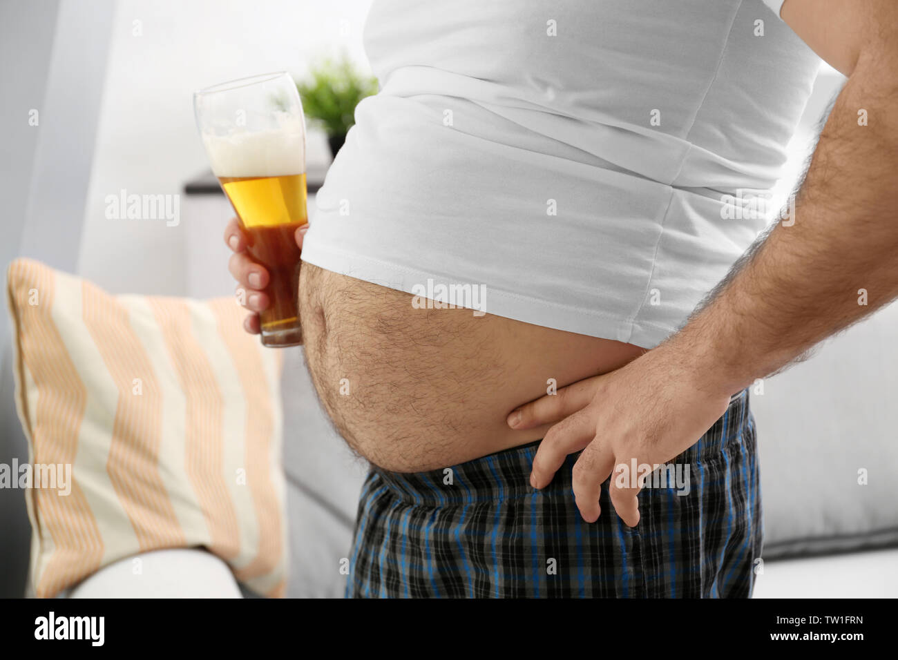 Uomo con grande pancia tenendo un bicchiere di birra in casa Foto Stock