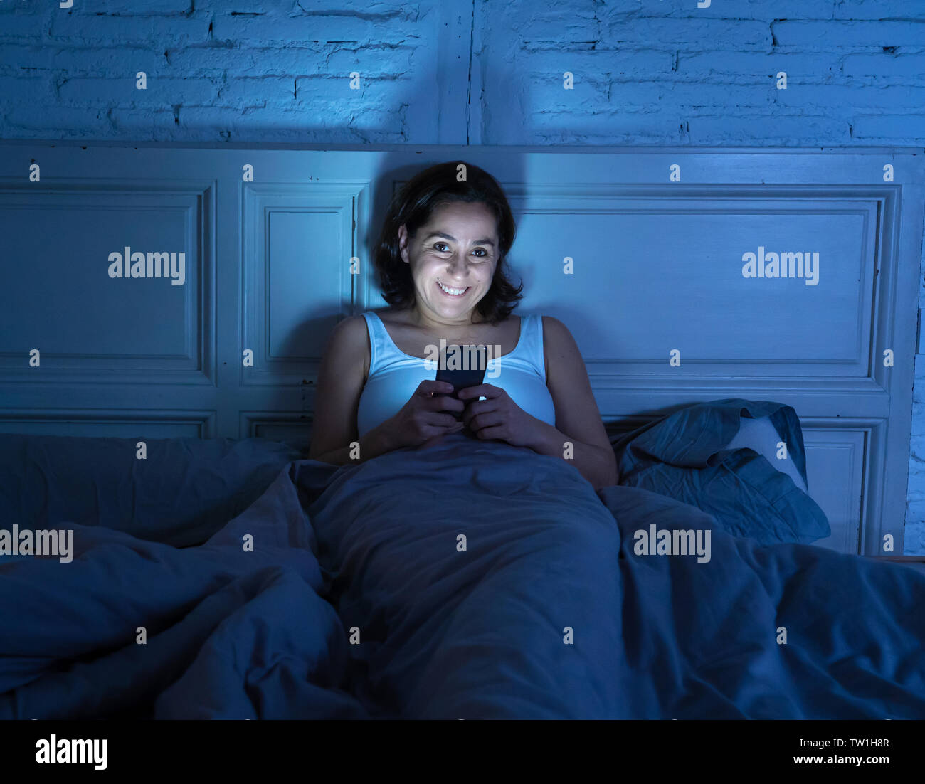 Donna ispanica svegli fino a tarda notte sulla smart phone a letto in una camera oscura. Uso di mobile per chattare e inviare messaggi in dipendenza da internet Foto Stock