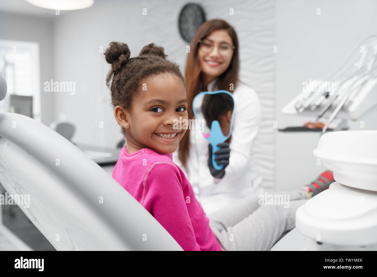 Poco ragazza africana seduta sulla poltrona del dentista guardando la fotocamera e sorridere mentre dentista femmina mantenendo specchio a backround. Bambino in visita dal dentista e godendo il sorriso dopo le procedure dentistiche. Foto Stock