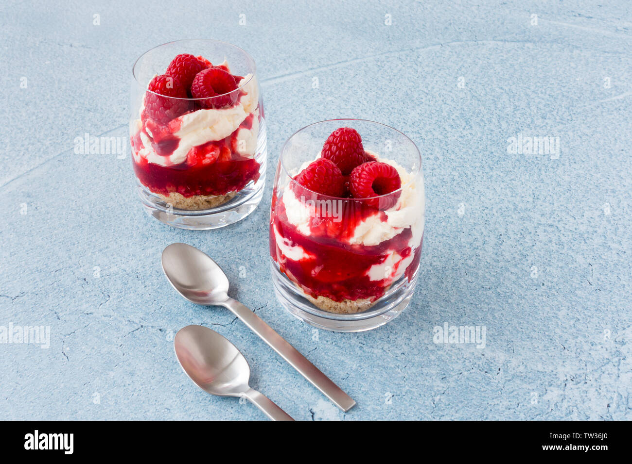 Lampone dolce con lamponi freschi, salsa di panna montata e i biscotti sminuzzati in bicchieri e cucchiai blu su sfondo di calcestruzzo con spazio di copia Foto Stock