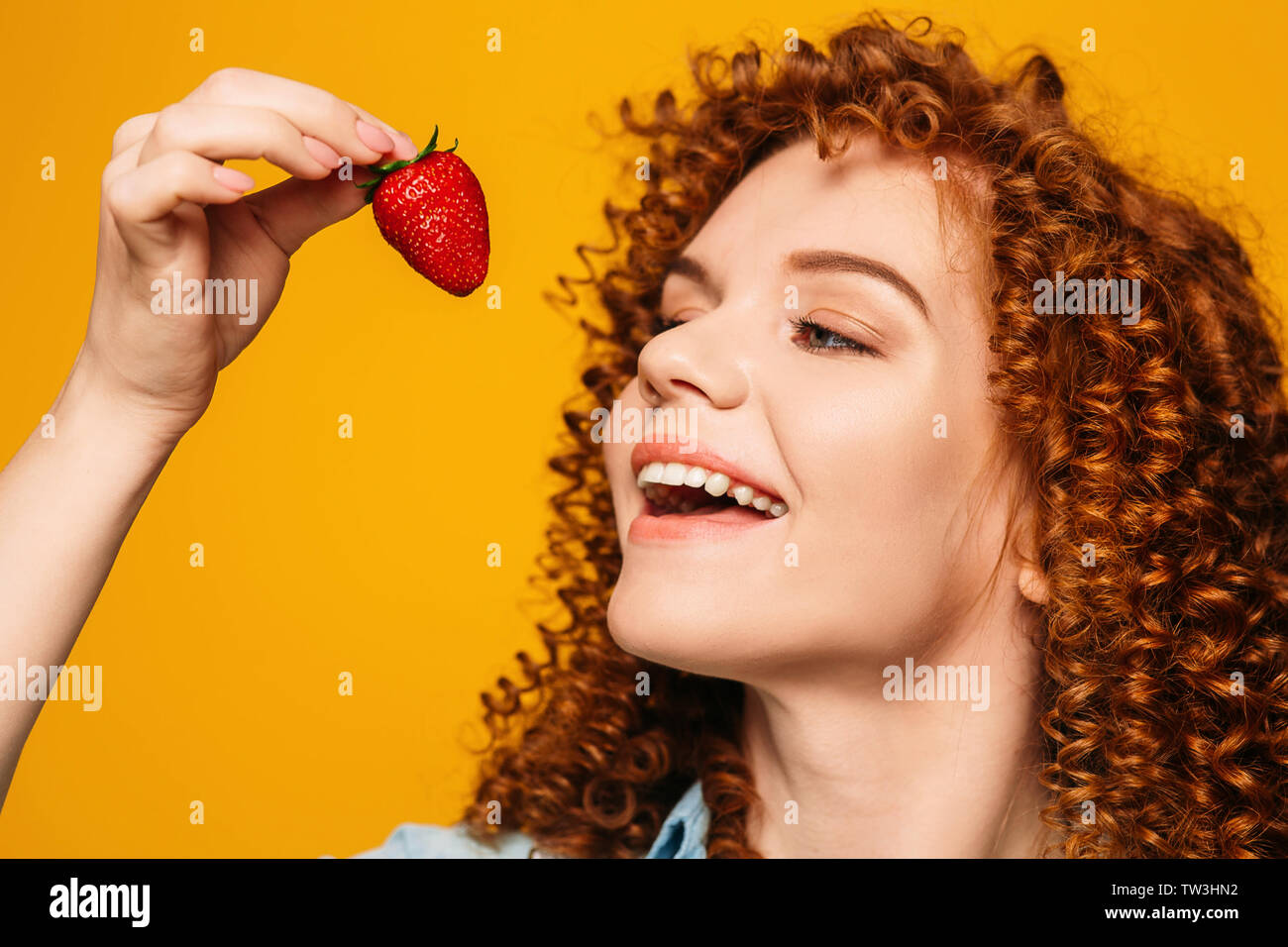 Ricci i capelli rossi donna con la bocca aperta vuole mangiare gustose fragole su sfondo giallo. Foto Stock