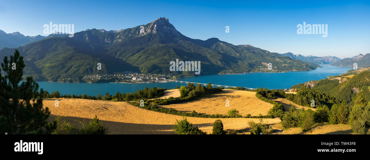 Panoramica del lago Serre-Poncon con il villaggio di Savines-le-Lac e campi di grano in estate la luce del mattino. Hautes-Alpes, Francia Foto Stock