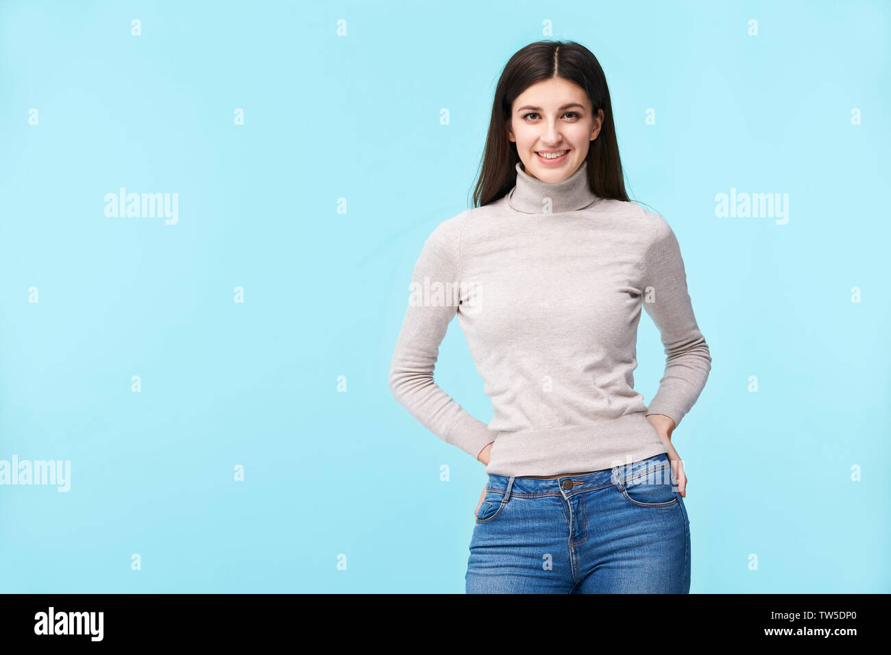 Ritratto di una giovane e bella donna caucasica, in piedi, mani sui fianchi, guardando la telecamera sorridendo, isolato su sfondo blu Foto Stock