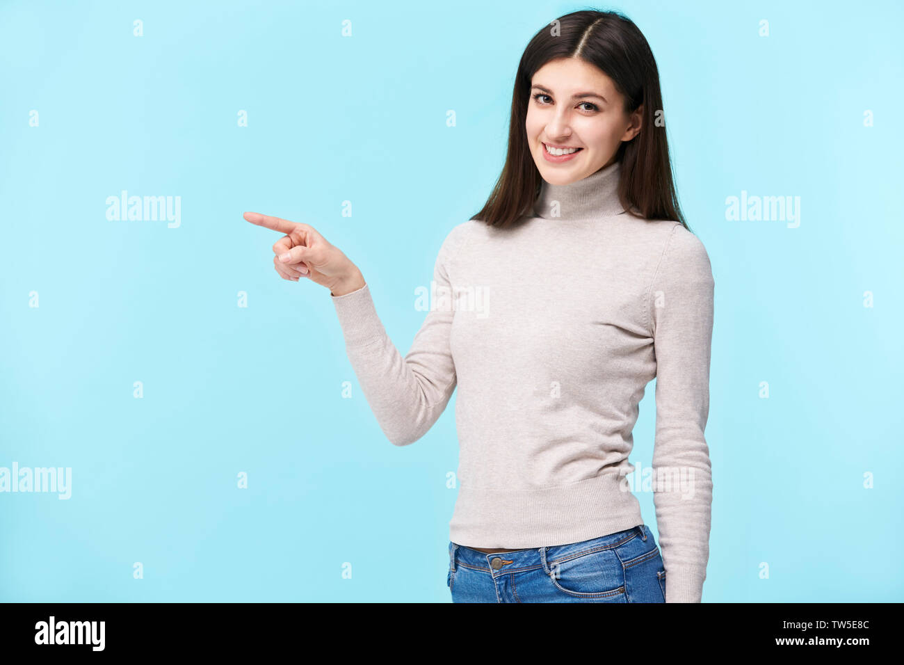 Ritratto di una giovane e bella donna caucasica, puntare il dito a qualcosa, guardando la telecamera sorridendo, isolato su sfondo blu Foto Stock