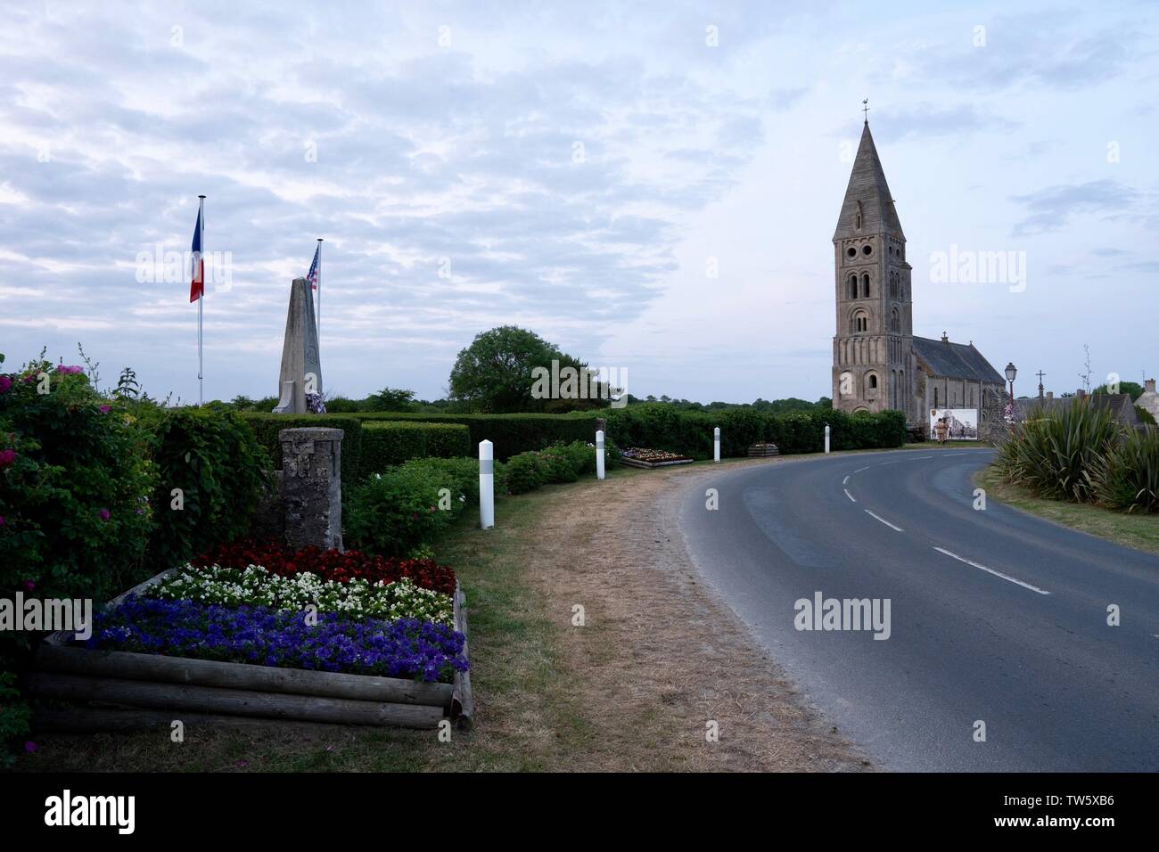 La chiesa all'entrata a Colleville sur Mer, un famoso villaggio francese, visto dal lato della strada - WW2 memorial e fiori in primo piano Foto Stock