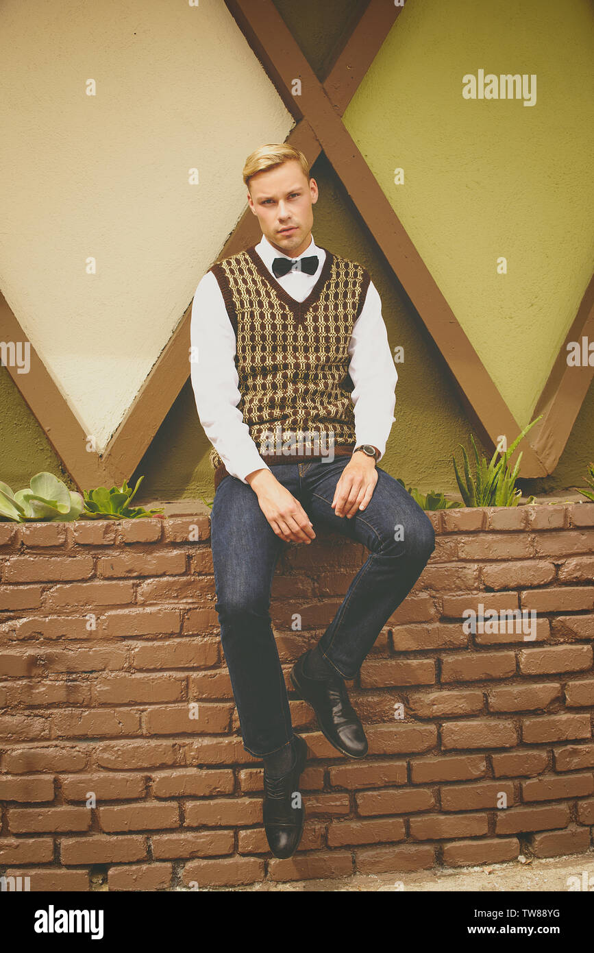 Bianco maschio caucasico si siede su un bordo di muro di mattoni, indossando un gilet, bow tie e jeans. Sembra rilassato. Un bel modello maschile. Foto Stock