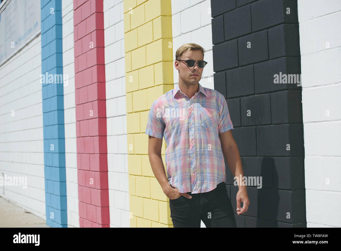Un modello maschio biondo che indossa una camicia a quadri e occhiali da sole occidentali, posando contro una parete blu in piedi all'aperto. Foto Stock