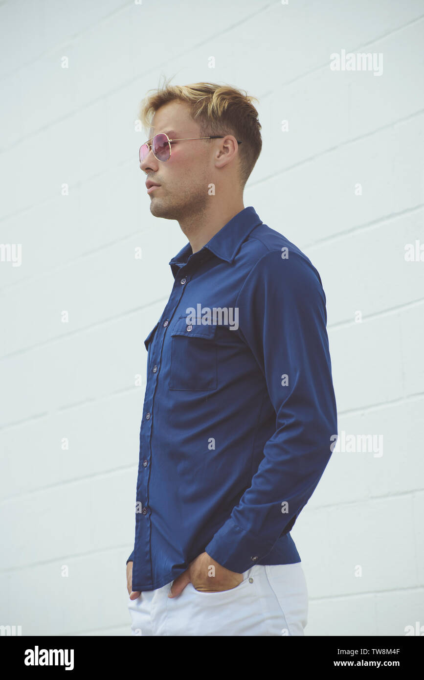 Un modello maschio caucasico biondo che si posa in una camicia d'epoca blu 70s, posando all'aperto su uno sfondo bianco parete. Un profilo laterale. Foto Stock