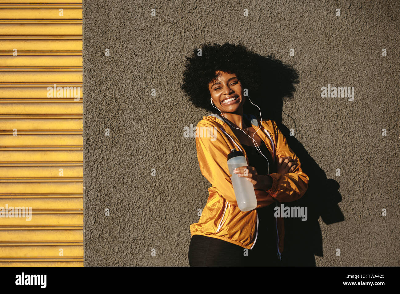 Fitness giovane donna in piedi all aperto con acqua in bottiglia e sorridente. Femminile in appoggio dopo allenamento, appoggiata ad una parete all'esterno. Foto Stock