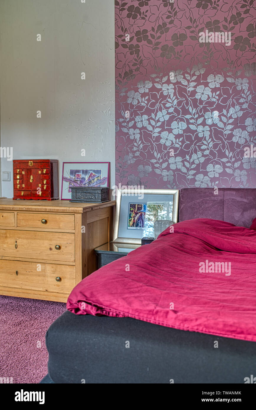 Dettagli della camera da letto con letto con materasso a molla e di una cassettiera, vari accessori e carta da parati viola Foto Stock