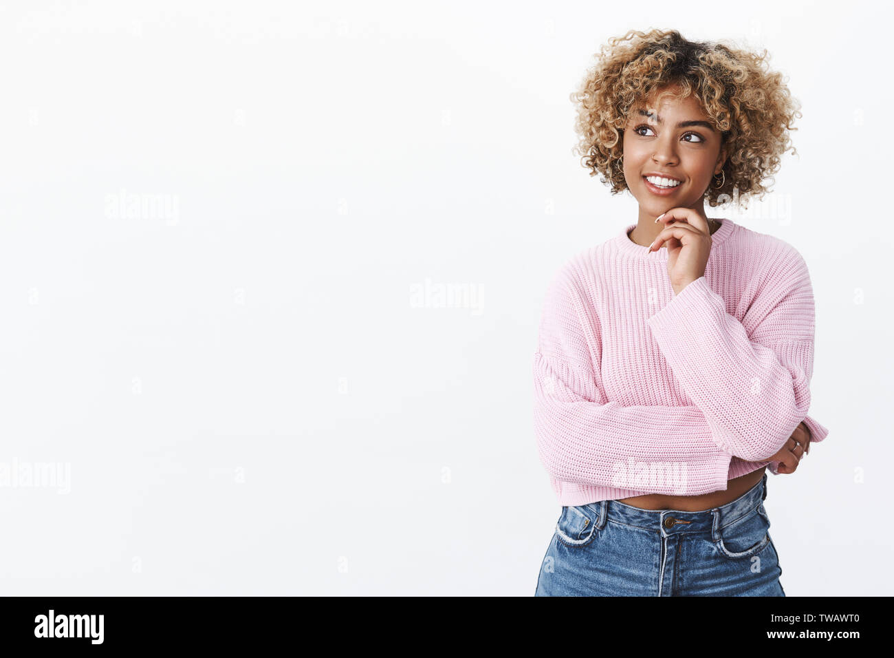 Attraente giovani afro-americano studente femmina con capelli biondi tenendo la mano sul mento e cercando il piacere ad angolo superiore sinistro come pensare, in posa Foto Stock
