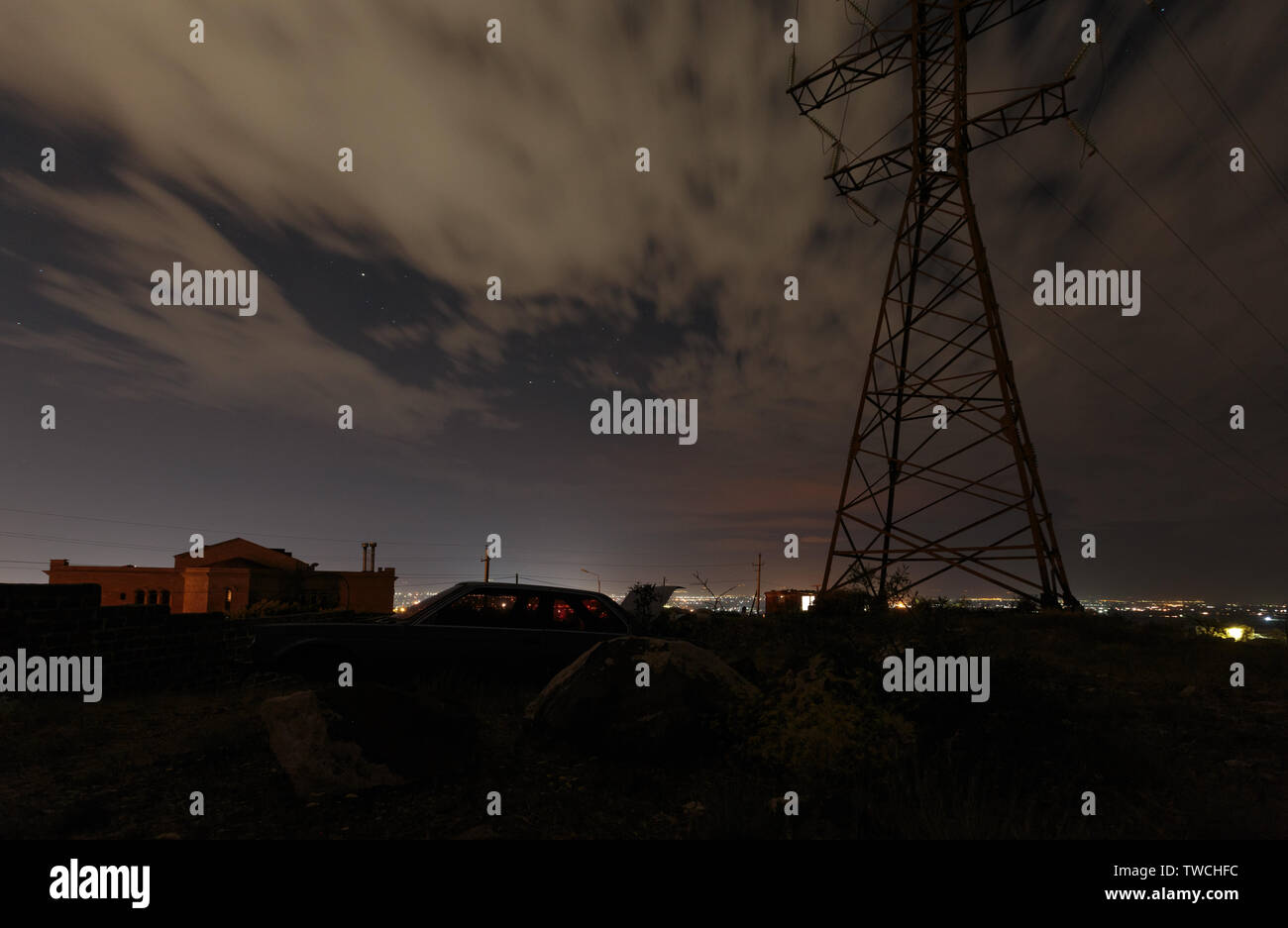 Supporto metallico per il cablaggio elettrico insieme con un broken auto contro lo sfondo delle luci della città di notte Foto Stock