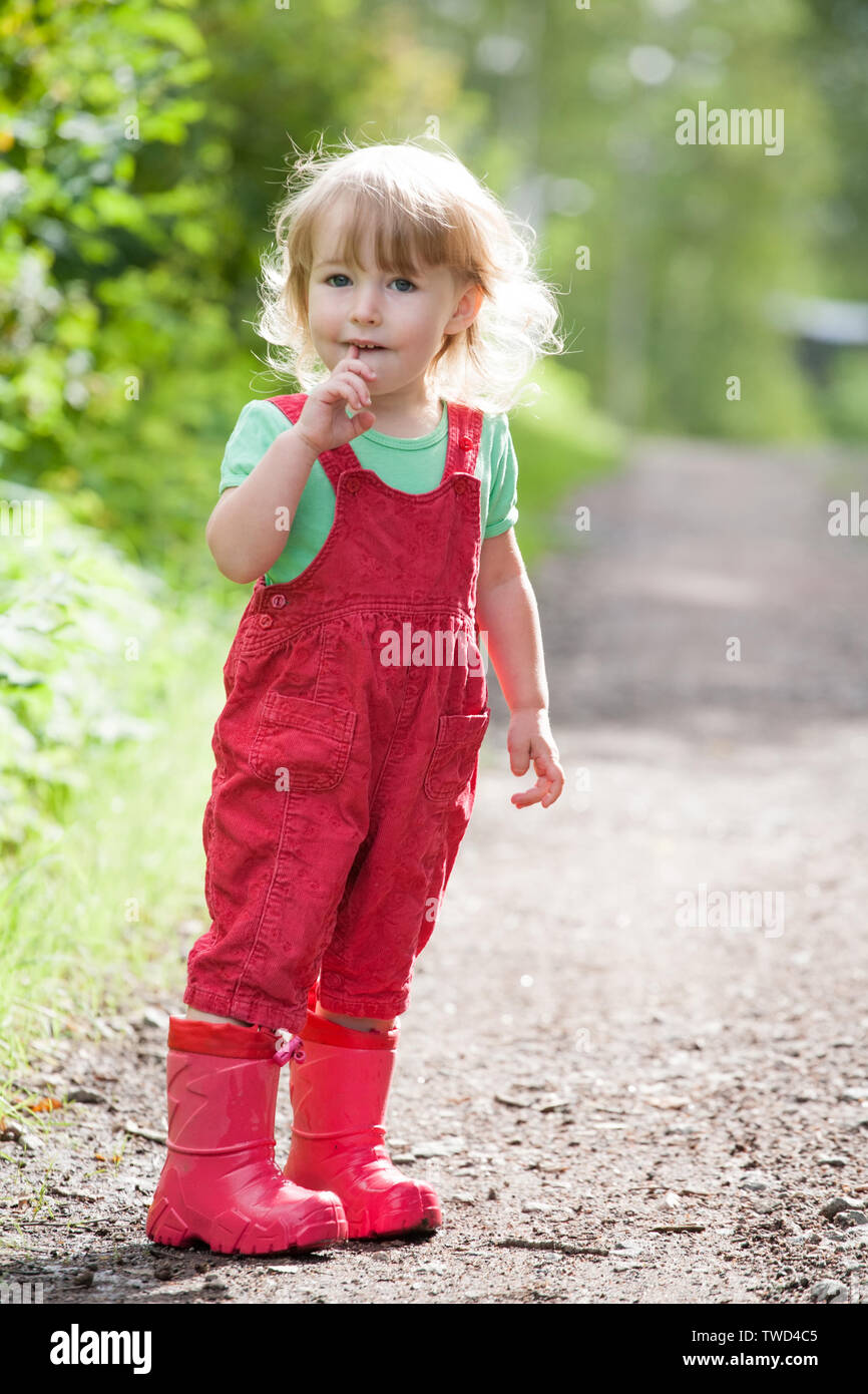 Funny bambina in rosso i pantaloni e stivali di gomma in piedi su estate sfondo all'aperto Foto Stock