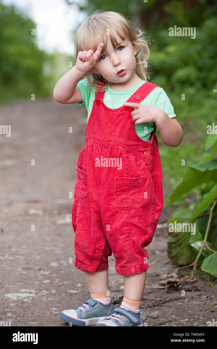 Carino due anni bionda ragazza bianco in pantaloni rossi full-size permanente sulla estate sfondo all'aperto Foto Stock