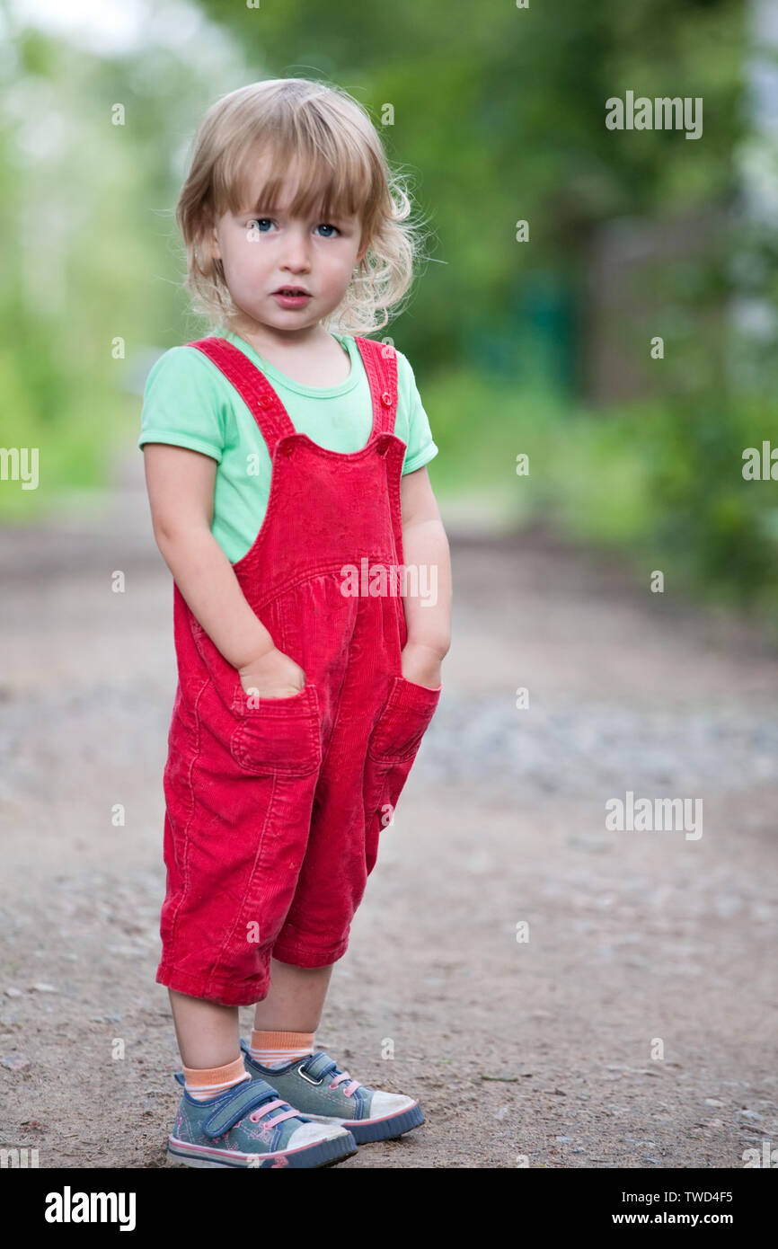 Carino due anni bionda ragazza bianco in pantaloni rossi full-size permanente sulla estate sfondo all'aperto Foto Stock