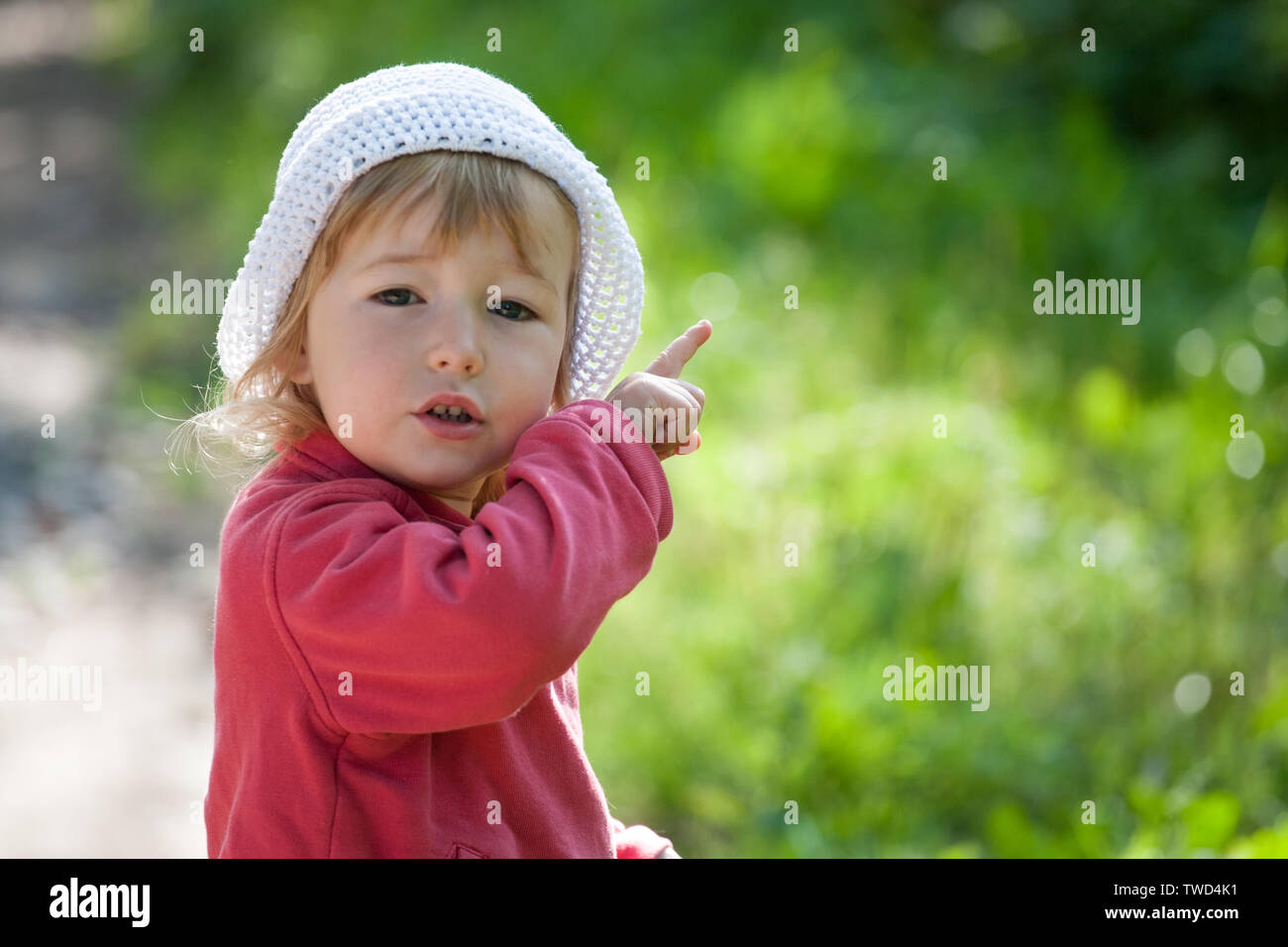 Bambino in giacca rossa e cappello bianco rivolto verso l'alto con il dito indice closeup vista sul verde sfondo all'aperto Foto Stock