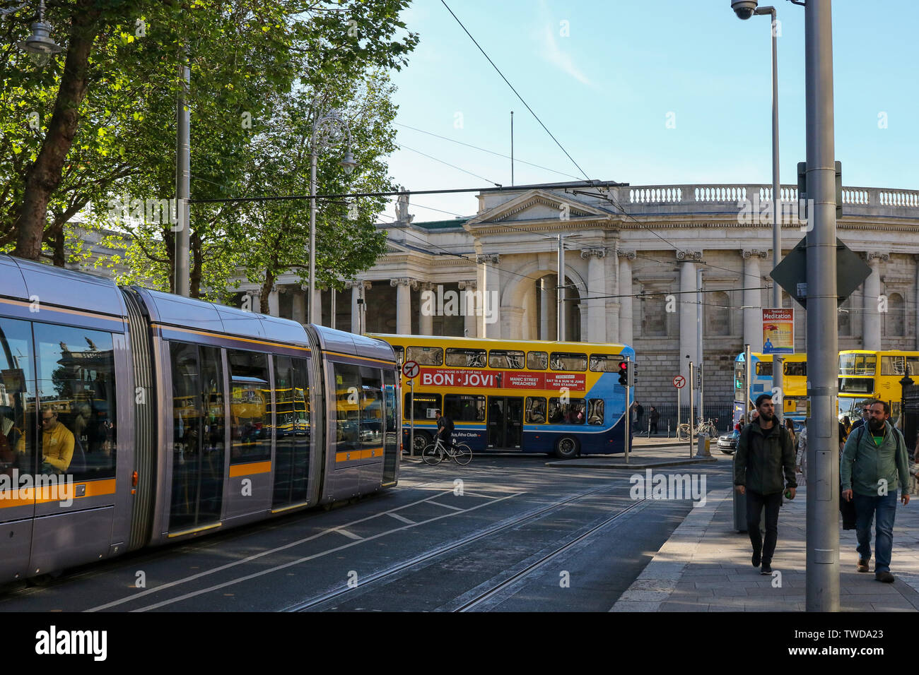 Scena stradale di Dublino, due uomini che camminano a Dublino in una serata estiva fuori dal Trinity College con un tram LUAS e autobus di Dublino sullo sfondo. Foto Stock