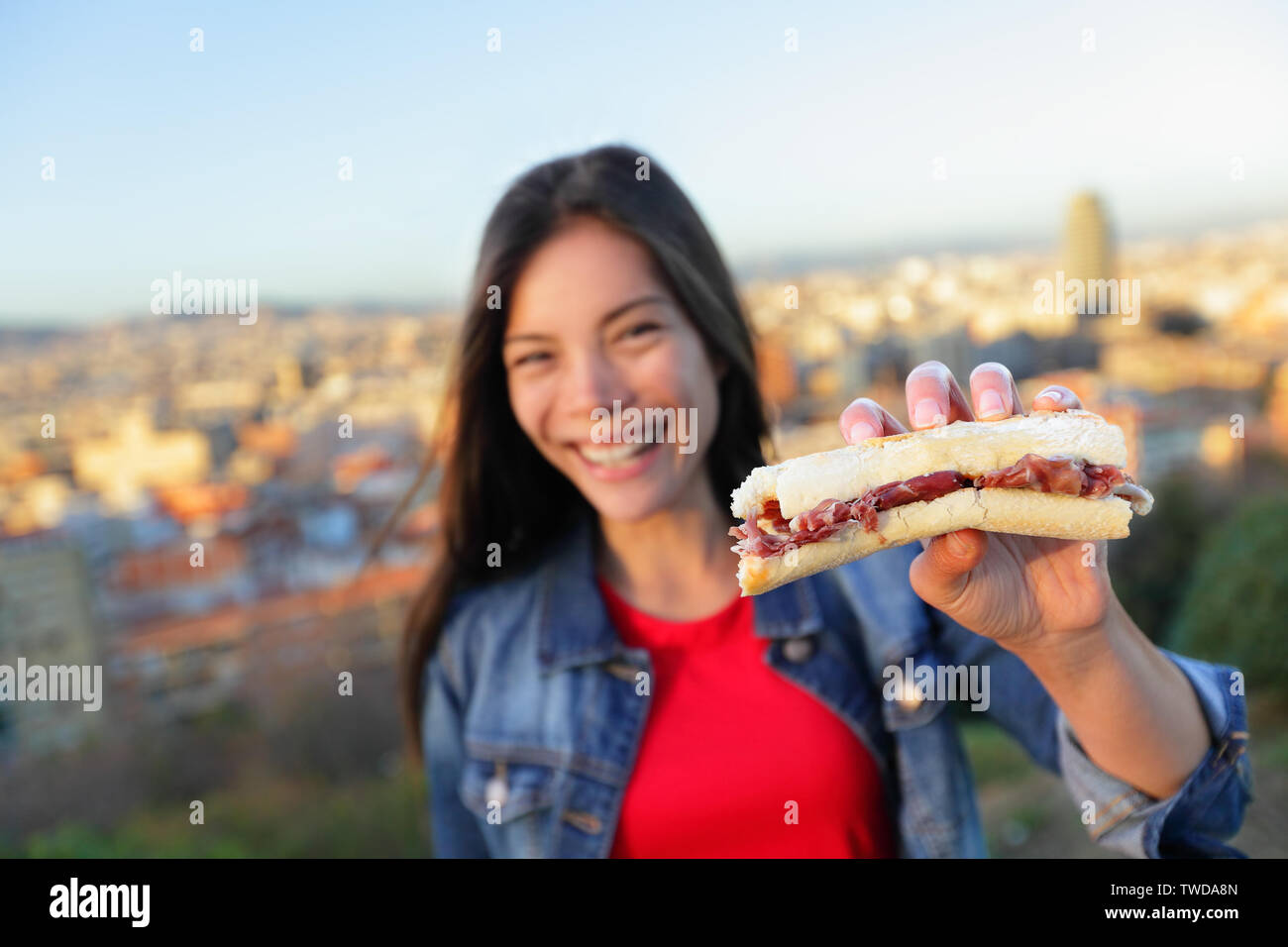 Jamon Iberico Sandwich. Donna di mangiare a Barcellona, Spagna mostra tradizionale cibo spagnolo, indurito il prosciutto Serrano. Focus su bocadillo, lo skyline di Barcellona in background. Foto Stock