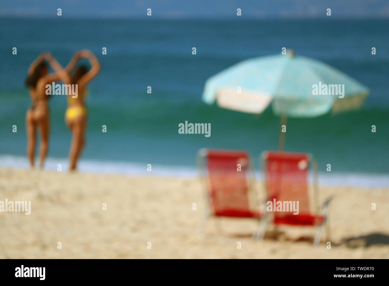 Immagine sfocata di una coppia di spiaggia rossa sedie e luce blu ombrellone sulla spiaggia sabbiosa contro vivid blue ocean Foto Stock