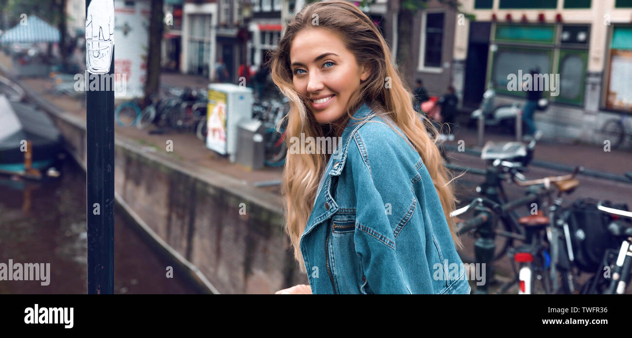 Ritratto di bella ragazza con ricci capelli biondi e largo sorriso. Deep Blue Eyes, luce specchio sul suo viso. Indossare il jeans giacca. In piedi vicino alla Foto Stock