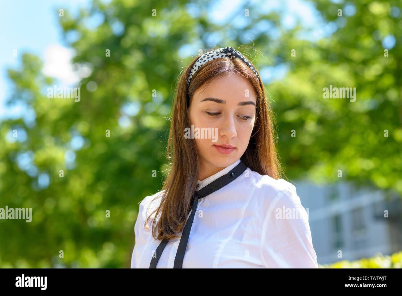 Sorridendo felice giovane donna con capelli lunghi marrone indossando un archetto e guardando verso il basso all'aperto sotto il sole Foto Stock