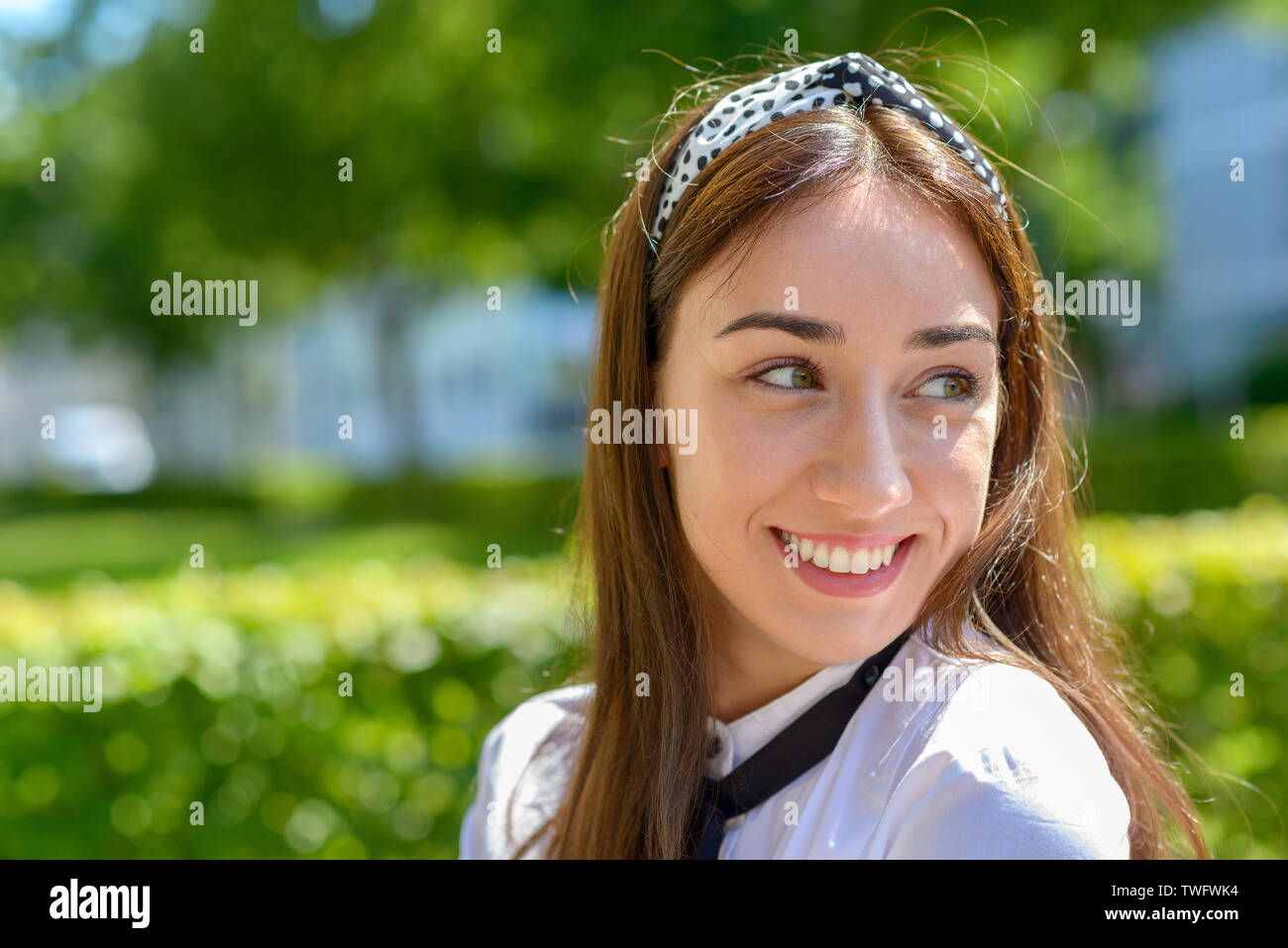 Sorridendo felice giovane donna con capelli lunghi marrone indossando un archetto e guardandosi le spalle all'aperto sotto il sole Foto Stock