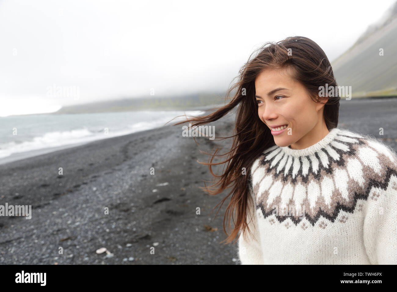 Bella donna camminando sulla spiaggia di sabbia nera su Islanda che indossa un maglione islandese. Piuttosto multirazziale modello femminile guardando pensieroso a mare oceano sorridendo felice. Foto Stock