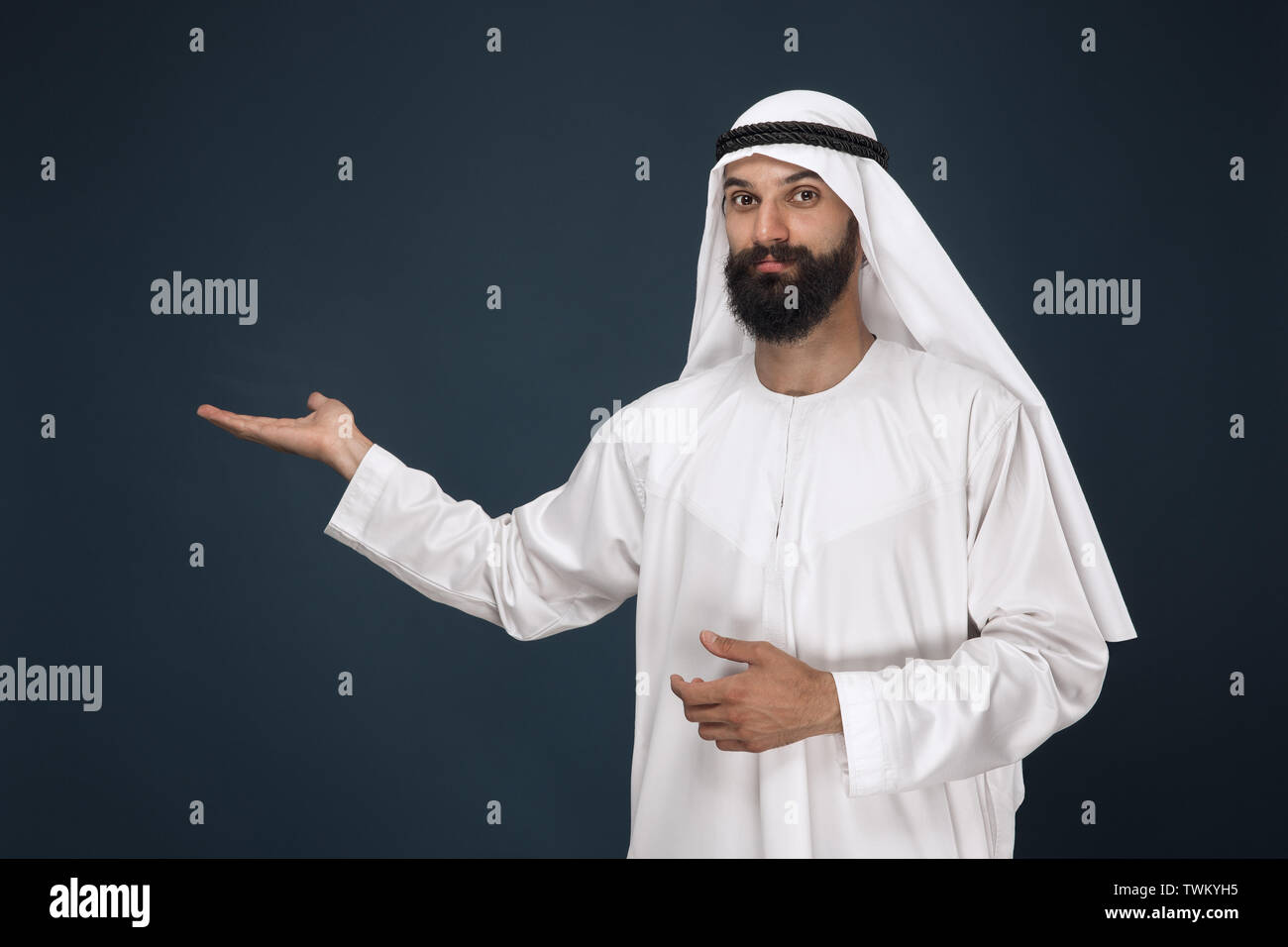 A mezza lunghezza Ritratto di arabian arabia uomo su blu scuro di sfondo per studio. Giovane maschio modello sorridente e puntamento. Concetto di business, finanza, espressione facciale, emozioni umane, tecnologie. Foto Stock