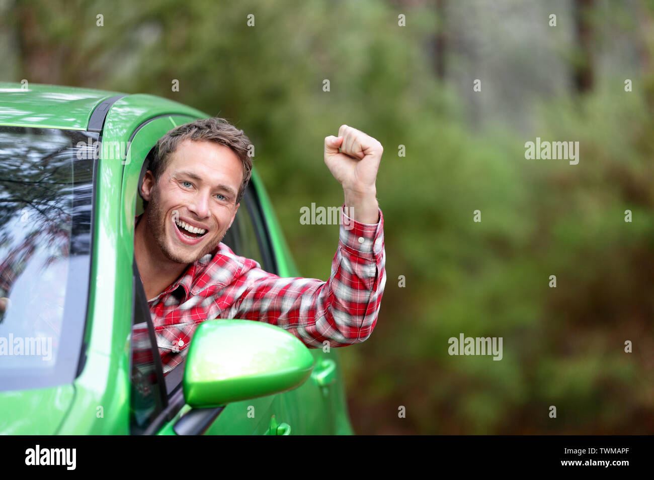 Energia verde biocarburanti auto elettrico conducente felice ed entusiasta. Uomo alla guida di un veicolo nuovo in allegro in natura foresta. Giovane maschio conducente guardando la fotocamera con il braccio sollevato il tifo. Foto Stock