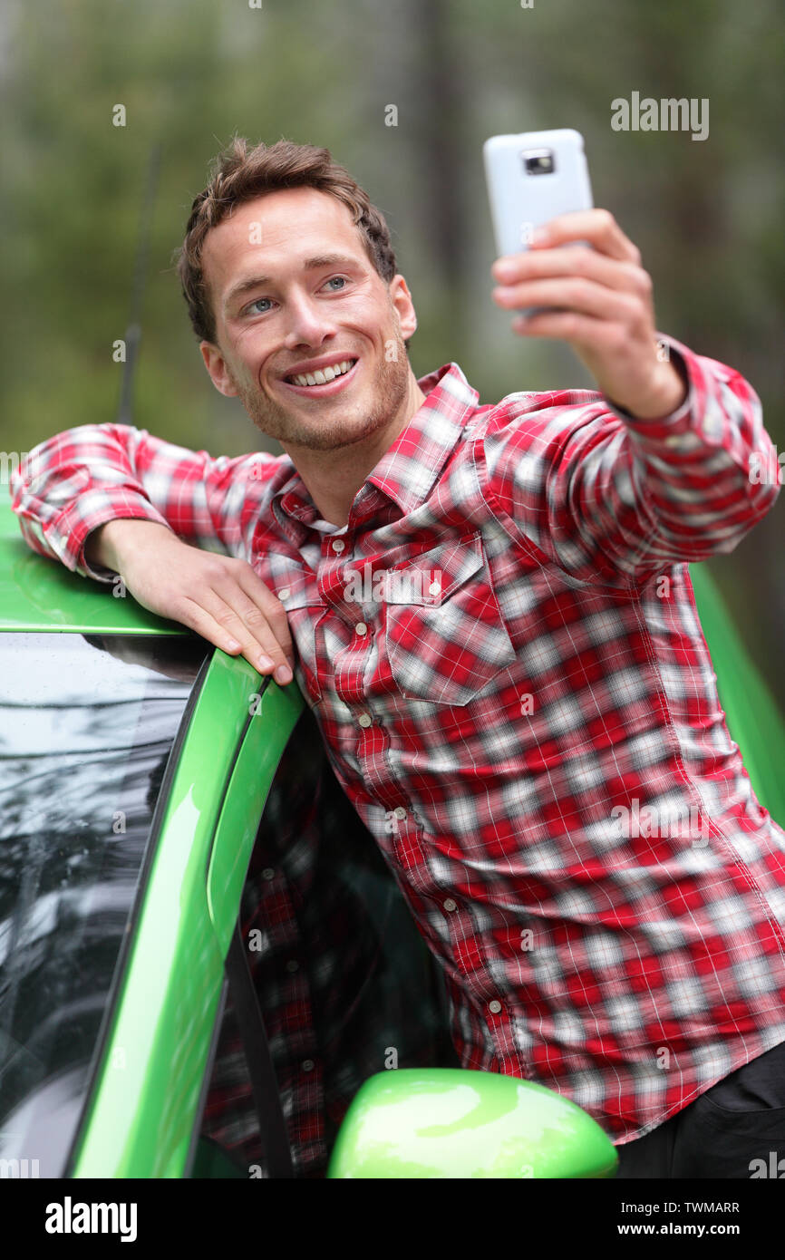 Autista con Auto prendendo selfie foto con lo smartphone dopo la guida nel nuovo verde auto. Uomo felice di scattare una foto con la fotocamera dello smartphone durante il viaggio viaggio su strada. Giovani caucasici modello maschio nel suo 20s. Foto Stock
