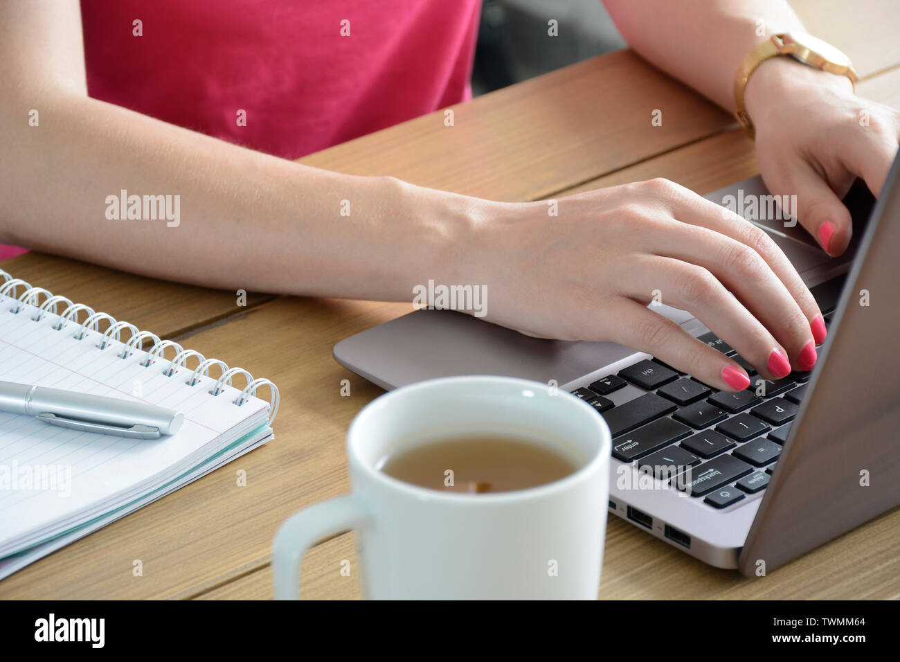 Giovane donna con una manicure rosa pulita utilizzando un moderno laptop. Con tazza di tè e taccuino. Foto Stock