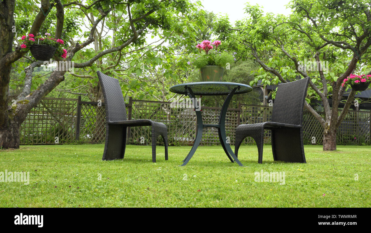 Due sedie di rattan e tavolo con vaso di fiori, sull'erba in un giardino estivo, circondato da alberi da frutto, coloratissimi cesti pendenti . Foto Stock