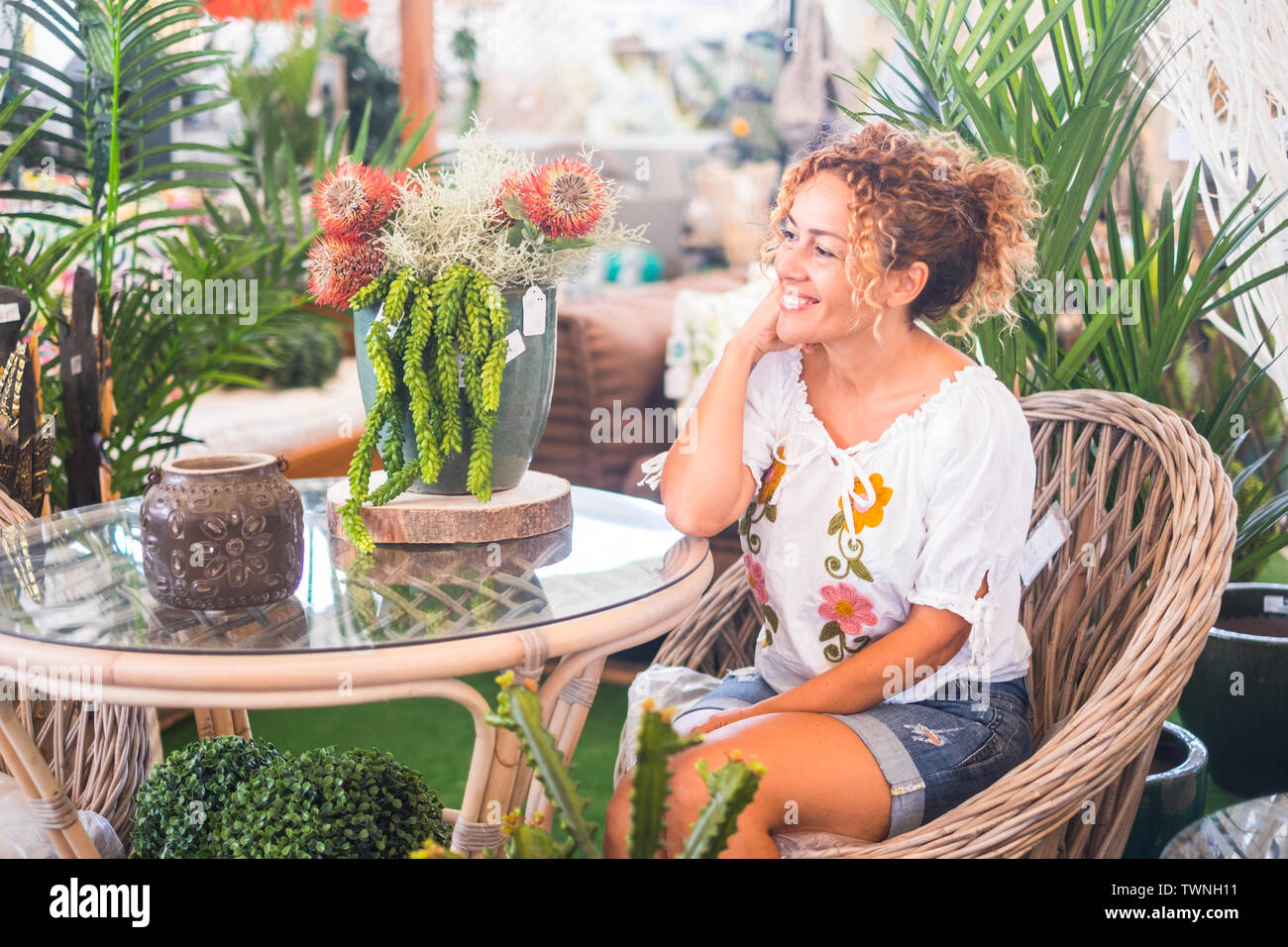 Giovane donna bionda con capelli ricci seduto su una sedia di vimini con molte piante si ammira la vegetazione. Il relax e il concetto di tempo libero per le persone enjoyin Foto Stock