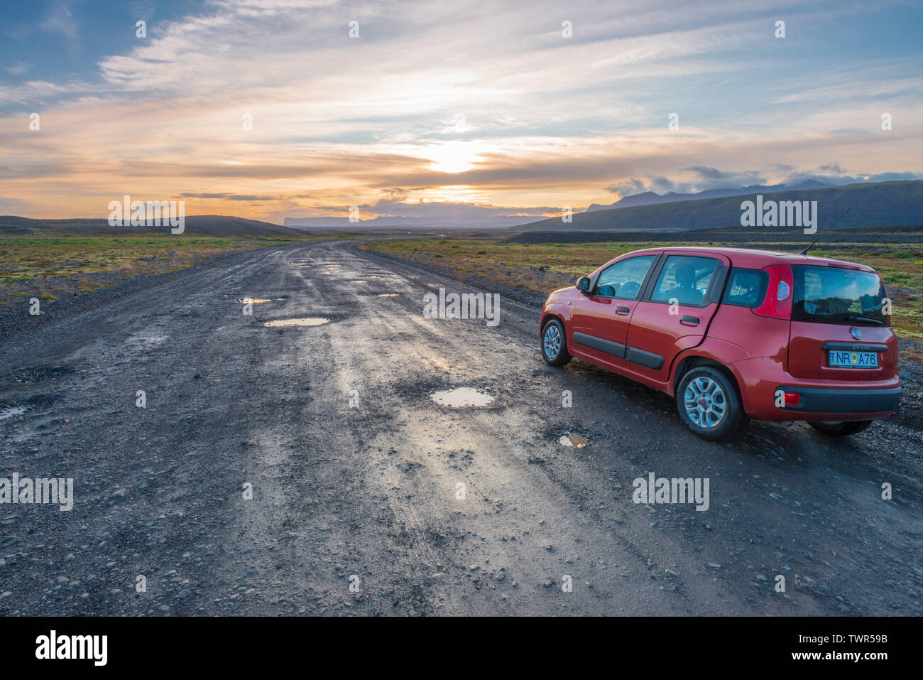 Tramonto sulla strada sterrata con buche in Islanda, Rosso Fiat Panda lungo la strada mentre il sole tramonta dietro le montagne. Foto Stock