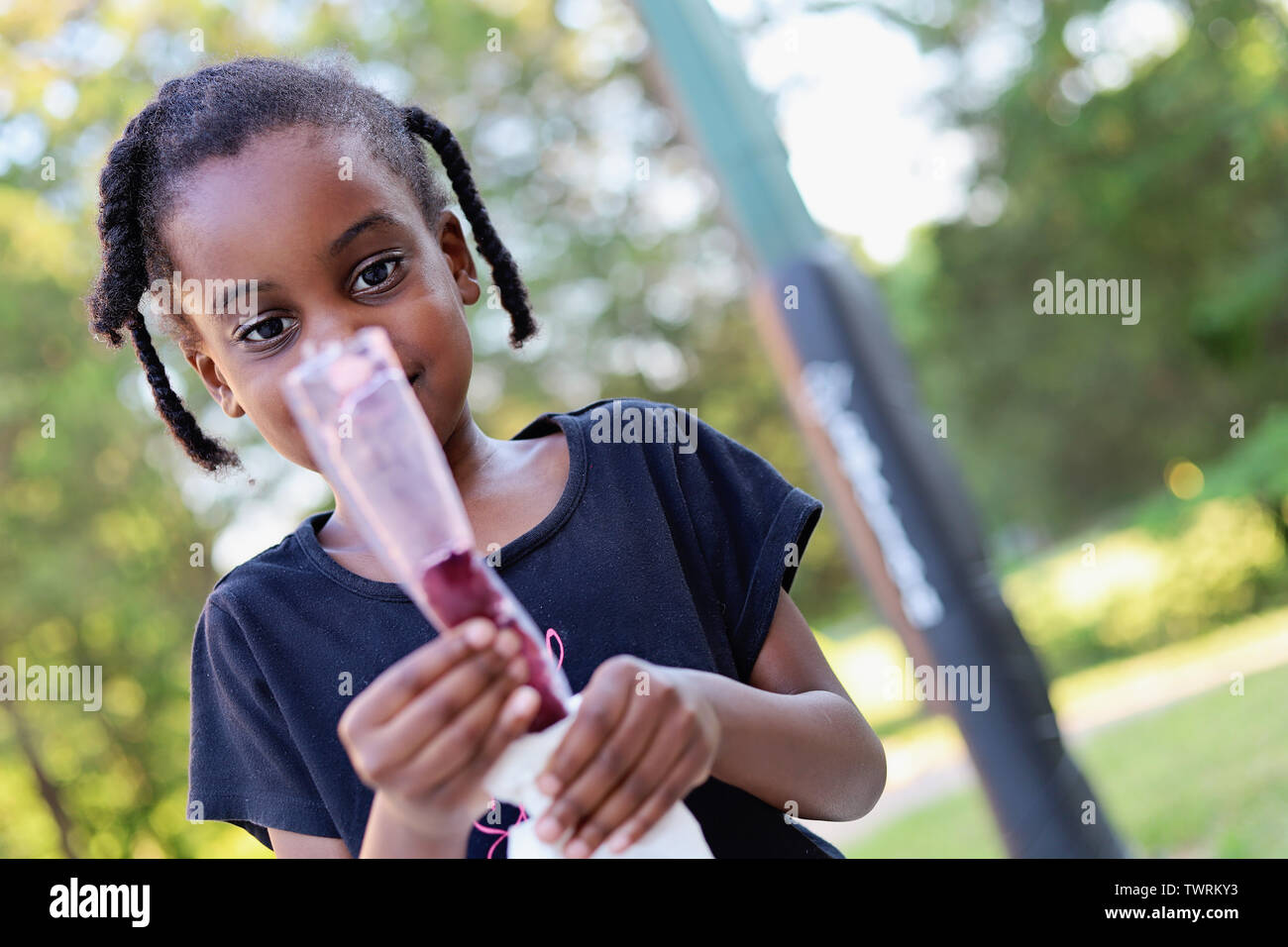 Little Black girl guardando un per lecca-lecca di ghiaccio su una soleggiata giornata estiva con alberi in background Foto Stock