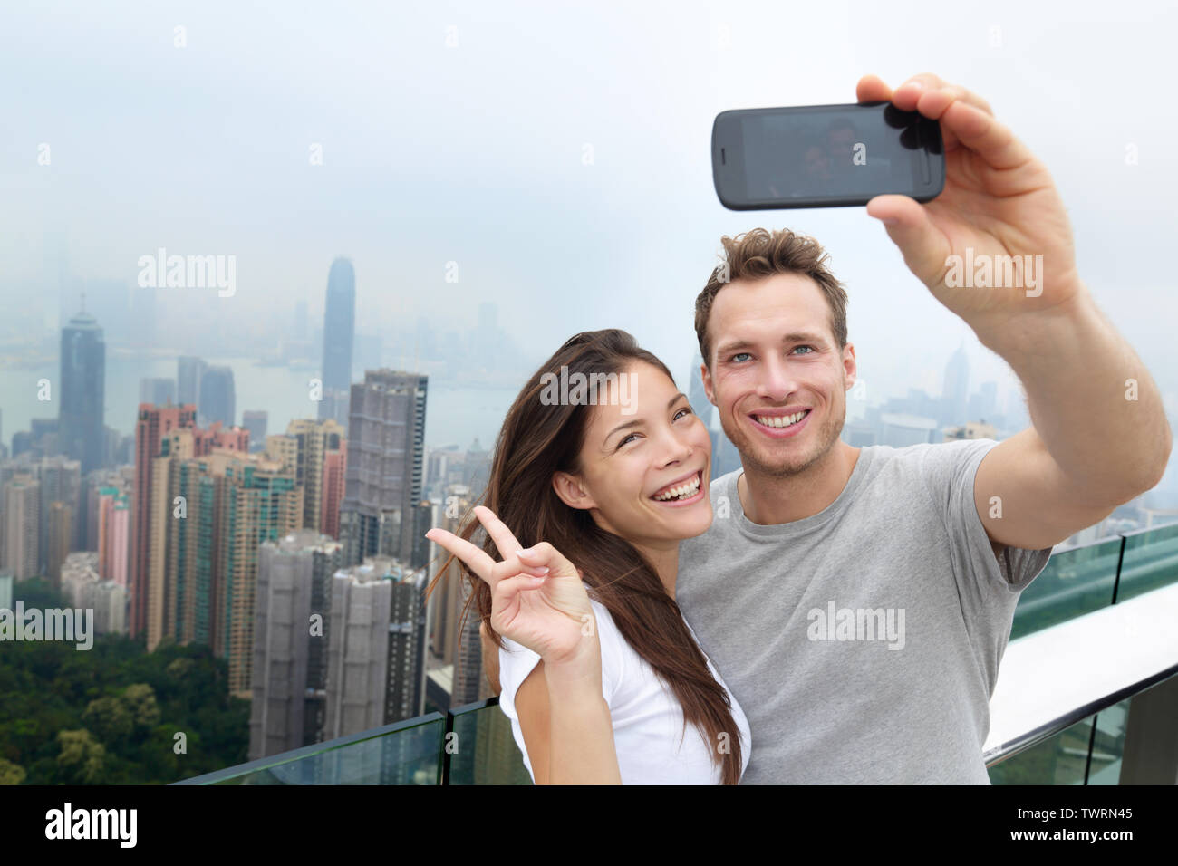 Hong Kong Victoria Peak turisti giovane tenendo selfie foto con lo smartphone gode di vista su Hong Kong e del porto di Victoria. Giovani multietnica felice coppia che viaggia in Asia. Foto Stock