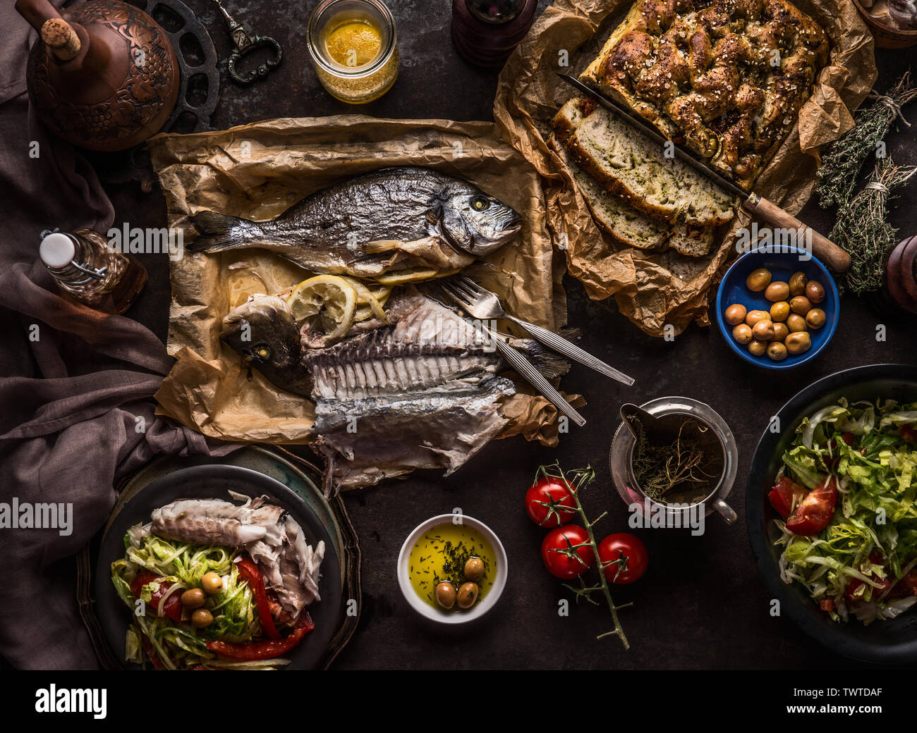 Cotta dorado pesce sulla tavola scuro con insalatiera , focaccia, pomodoro e olive, vista dall'alto. Mediterraneo il pranzo o la cena. Il cibo italiano concept Foto Stock
