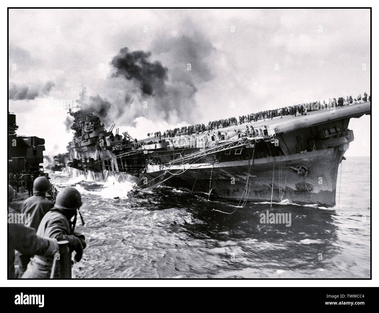 WW2 immagine della portaerei USS Franklin (CV-13) attaccato nel Pacifico durante la II Guerra Mondiale, Marzo 19, 1945. Fotografata da PHC Albert giovenco dall'incrociatore USS Santa Fe (CL-60), che è stata a fianco di assistere con mezzi antincendio e di soccorso. Il supporto è sul fuoco e la quotazione dopo che essa è stata colpita da un giapponese di attacco aereo durante il funzionamento al largo delle coste del Giappone - l'equipaggio è chiaramente visibile sul ponte di volo. Dopo l'attacco la nave giacevano morti nell'acqua, ha preso un 13° elenco di dritta, perso tutte le comunicazioni radio e arrostito sotto il calore da incendi avvolgente. Foto Stock