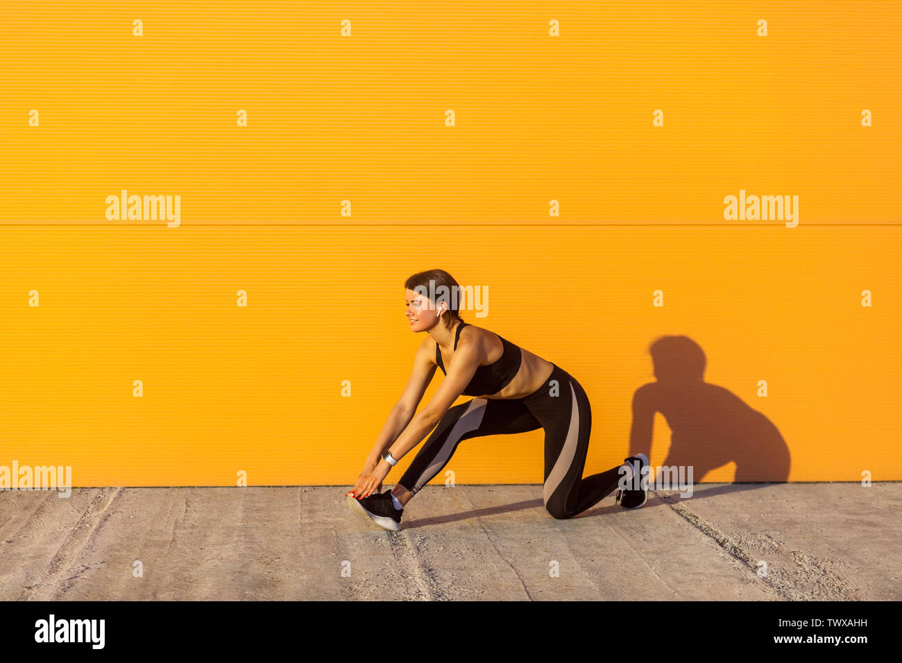 Vista laterale del giovane bella sportiva femmina yogi in nero sportwear stiramento sulla strada a pavimento sul ginocchio, guardando dritto, Outdoor, arancione Foto Stock