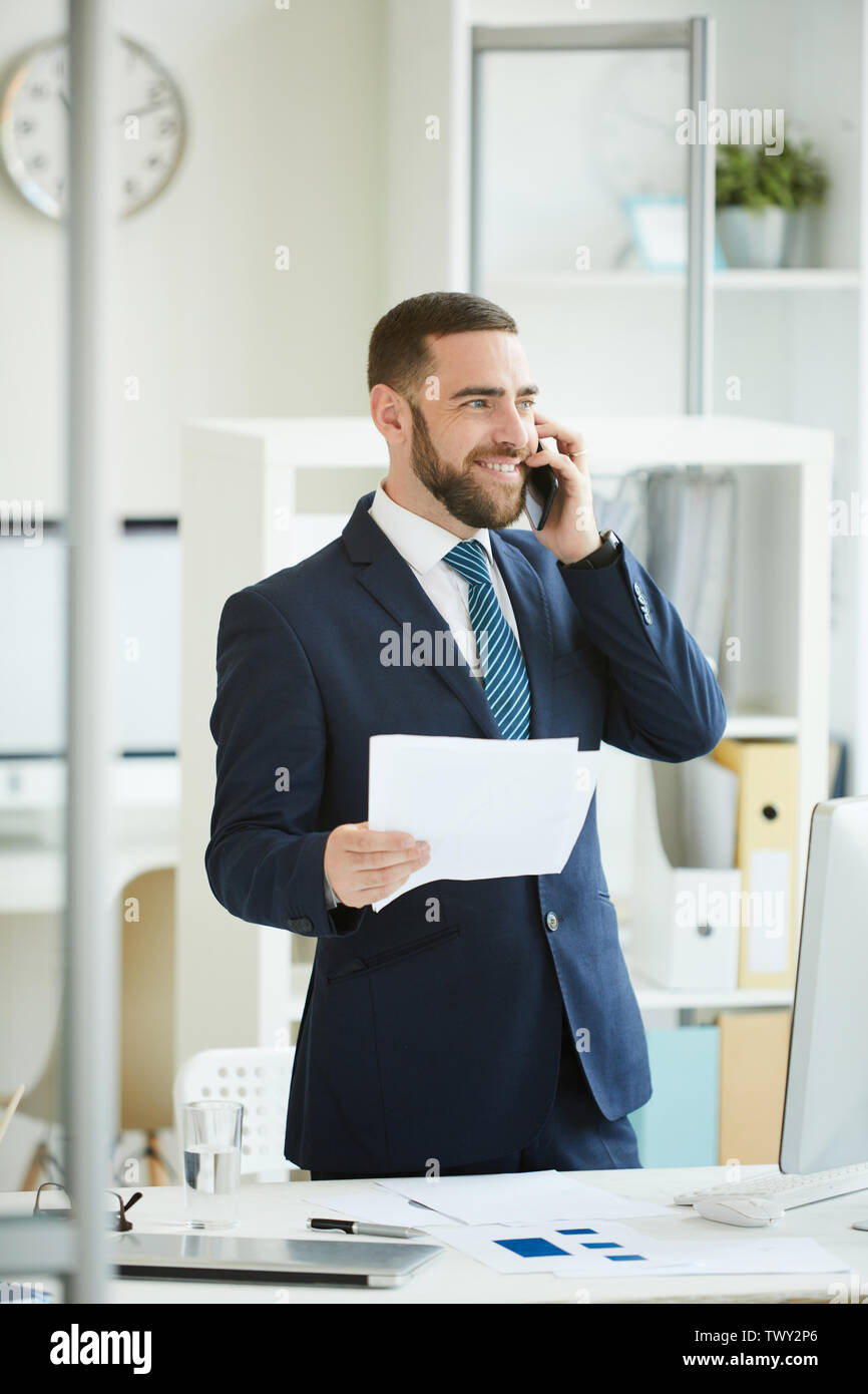 Ottimista bel giovane imprenditore barbuto in blu scuro tuta in piedi in un ufficio moderno ed avente conversazione telefonica mentre discutete le carte con c Foto Stock