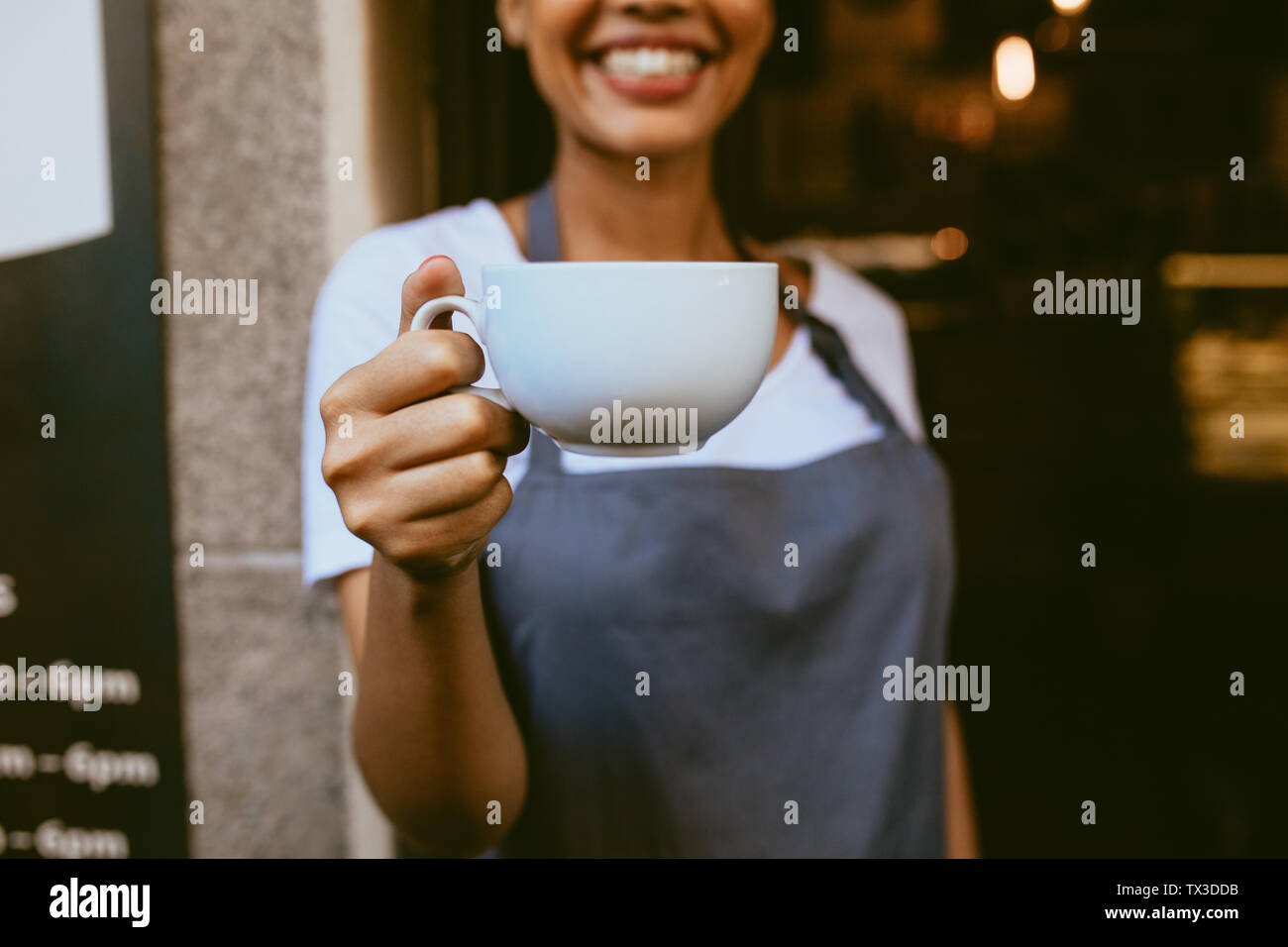 Close up femminile di barista che offre una tazza di caffè mentre Standing at the coffeeshop porta. Focus sulla tazza di caffè in mano di una femmina di cafe proprietario. Foto Stock