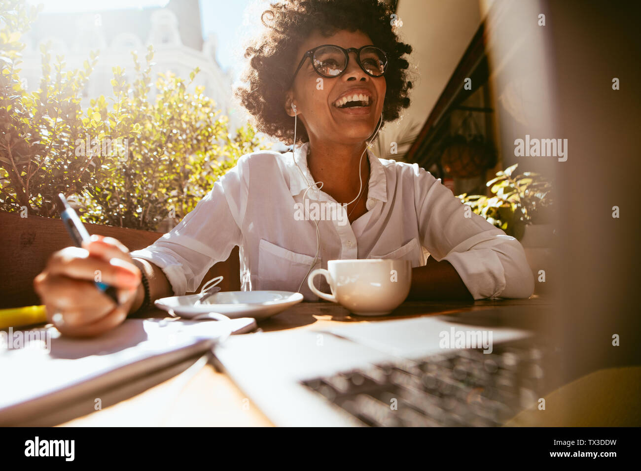 Felice giovane donna africana la scrittura di note durante la seduta a una caffetteria con un computer portatile sul tavolo. Lavoro femminile da un coffee shop. Foto Stock